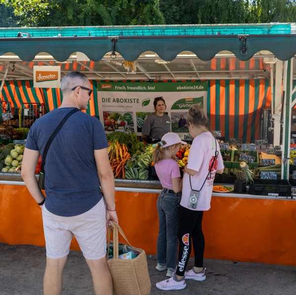 KI generiert: Marktstand mit Bio-Gemüse. Menschen schauen das Angebot an. Text: "DIE ABOKISTE" und "abokiste24.de".