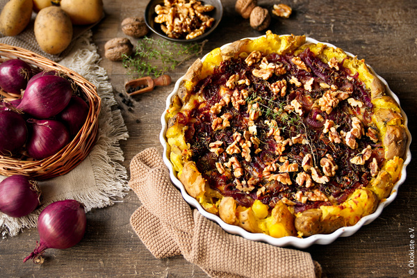 KI generiert: Das Bild zeigt eine appetitliche Tarte, die mit roten Zwiebeln, Walnüssen und Kräutern belegt ist. Daneben liegen Zutaten wie rote Zwiebeln und Kartoffeln auf einem rustikalen Holztisch.
