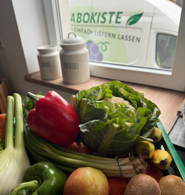 KI generiert: Eine Kiste mit frischem Gemüse und Obst vor einem Fenster. Text: "ABOKISTE".