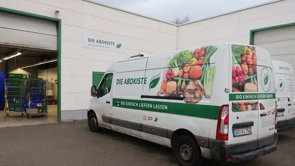 KI generiert: Transporter vor einem Gebäude mit der Aufschrift "DIE ABOKISTE - BIO EINFACH LIEFERN LASSEN".