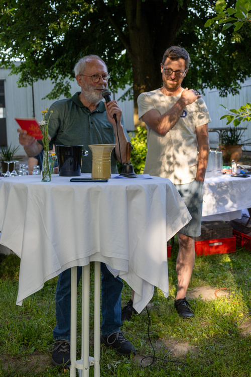 KI generiert: Zwei Männer stehen an einem Tisch im Freien. Einer hält ein Mikrofon.