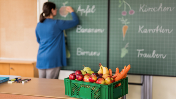 KI generiert: Eine Person schreibt Früchtenamen an eine Tafel, vorne steht ein Obstkorb. Text: "Apfel, Bananen, Birnen, Kirschen, Karotten, Trauben".