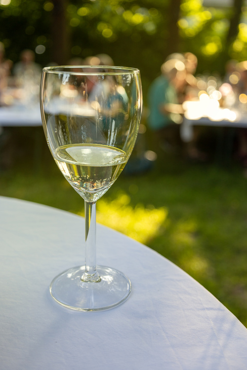 KI generiert: Weinglas auf Tisch im Vordergrund, verschwommen Menschen im Hintergrund.