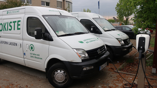 KI generiert: Zwei Lieferwagen mit der Aufschrift "Ökokiste" laden an einer Ladestation. Text: "Ich fahre mit 100% Ökostrom".