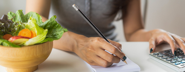 KI generiert: Eine Person macht Notizen mit einem Bleistift neben einer Schüssel Salat und verwendet einen Taschenrechner.