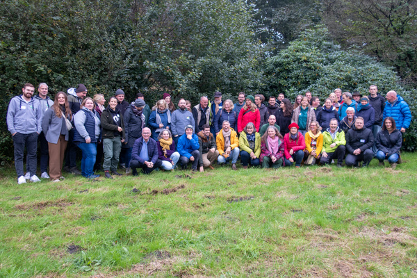 KI generiert: Das Bild zeigt eine große Gruppe von Menschen, die draußen auf einer Wiese zusammenstehen und in die Kamera lächeln. Sie sind von Bäumen und Büschen umgeben und scheinen an einem Gruppen- oder Gemeinschaftstreffen teilzunehmen.