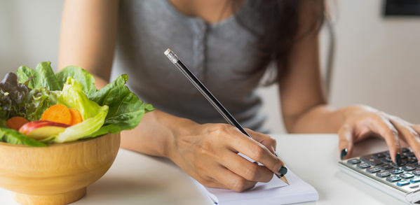 KI generiert: Frau schreibt mit einem Bleistift in ein Notizbuch neben einer Schüssel Salat und einem Taschenrechner.