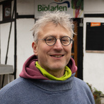 KI generiert: Ein lächelnder Mann mit Brille steht vor einem Fachwerkgebäude. Im Hintergrund ist ein Schild mit der Aufschrift "Bioladen" zu sehen.