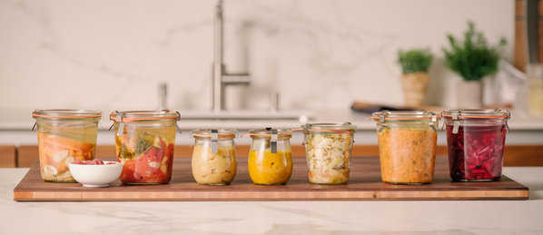 Verschiedene Einmachgläser mit buntem, eingelegtem Gemüse auf einem Holzbrett in einer Küche.